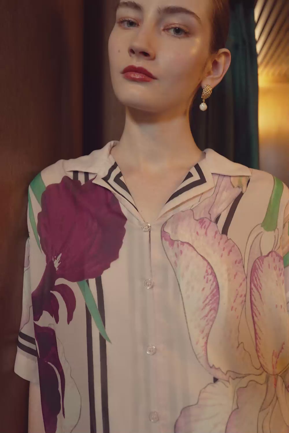 Model wearing BEHOLD unisex Cuban Shirt and paperbag palazzo pants in Iris floral print by Reckless Ericka, featuring oversized blooms and relaxed tailoring. Filmed in a moody, warm-lit indoor setting.