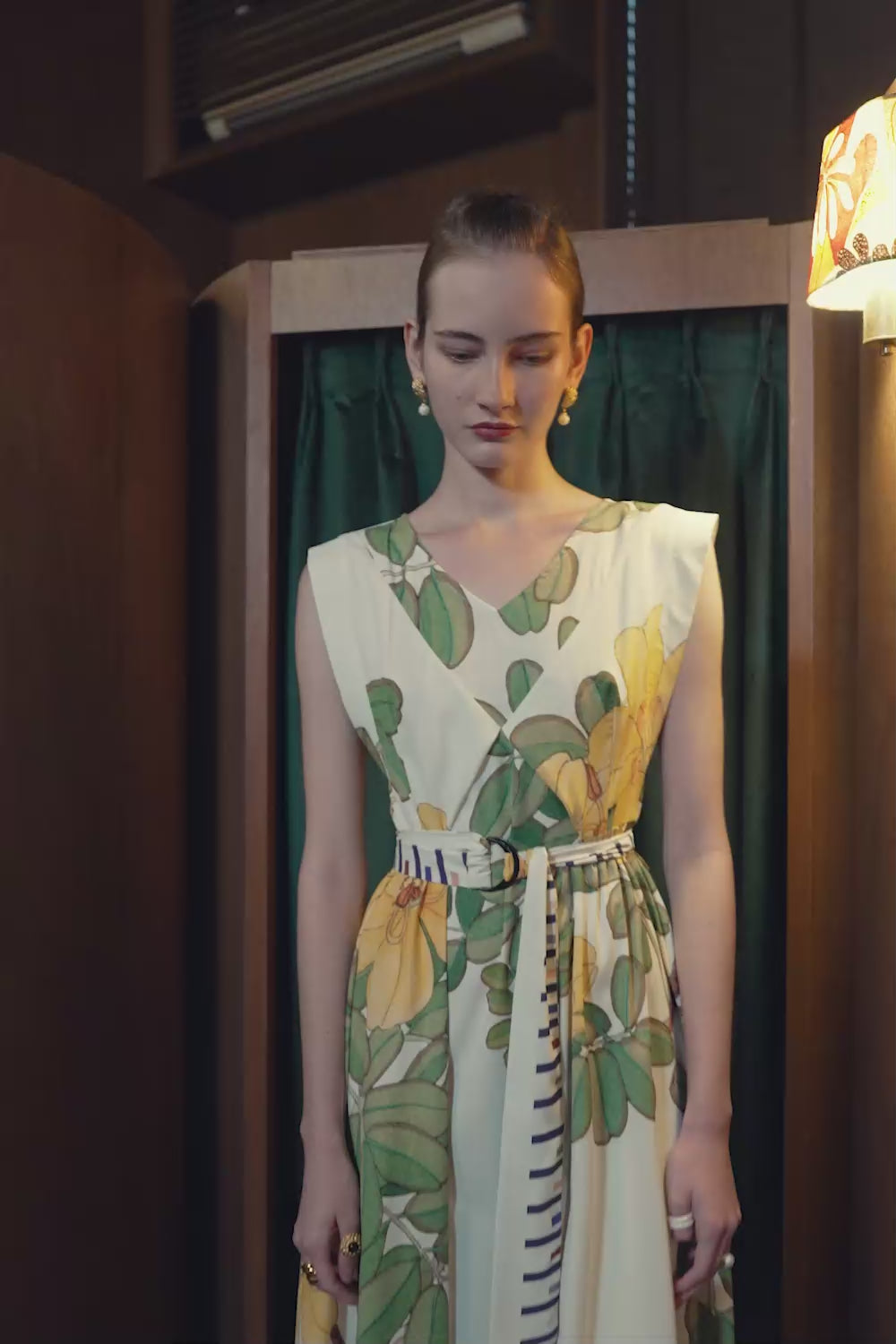 Model walking and posing in BEHOLD Origami Square Sleeve Dress with yellow Allamanda floral print, styled with striped sash, captured in an indoor setting.
