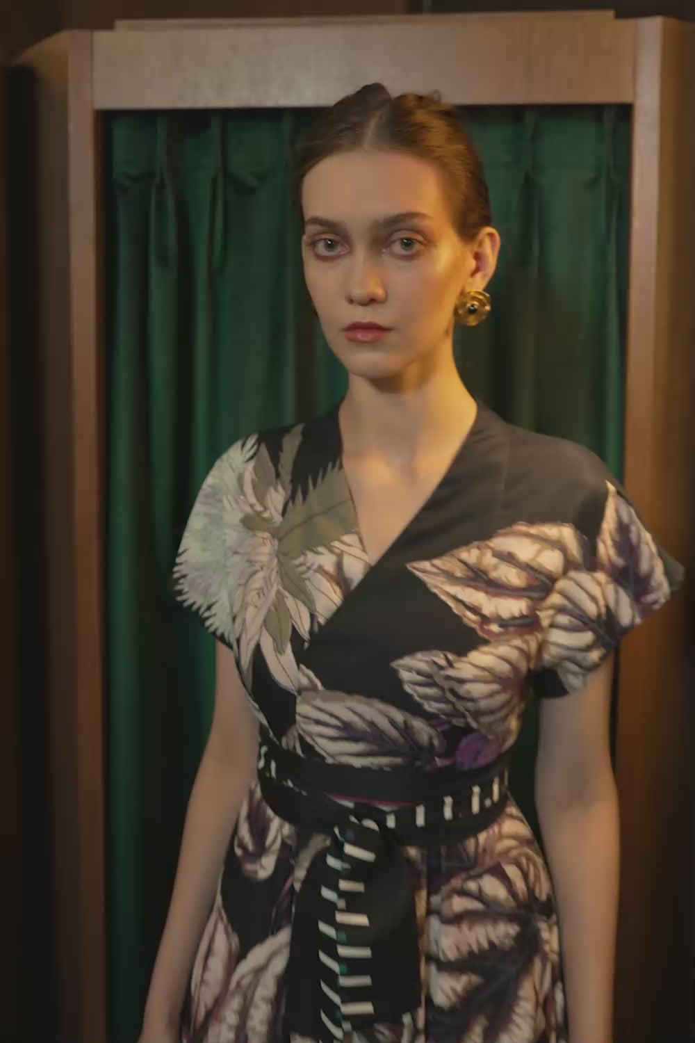 Model walking in the BEHOLD Blended Collar Wrap Midi Dress featuring oversized botanical prints, a blended Mandarin-meets-V neckline, and a obi belt. Filmed in a wooden-paneled room with a green curtain backdrop.