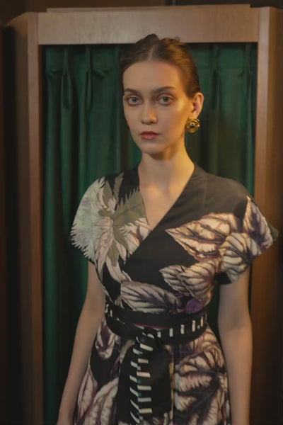 Model walking in the BEHOLD Blended Collar Wrap Midi Dress featuring oversized botanical prints, a blended Mandarin-meets-V neckline, and a obi belt. Filmed in a wooden-paneled room with a green curtain backdrop.
