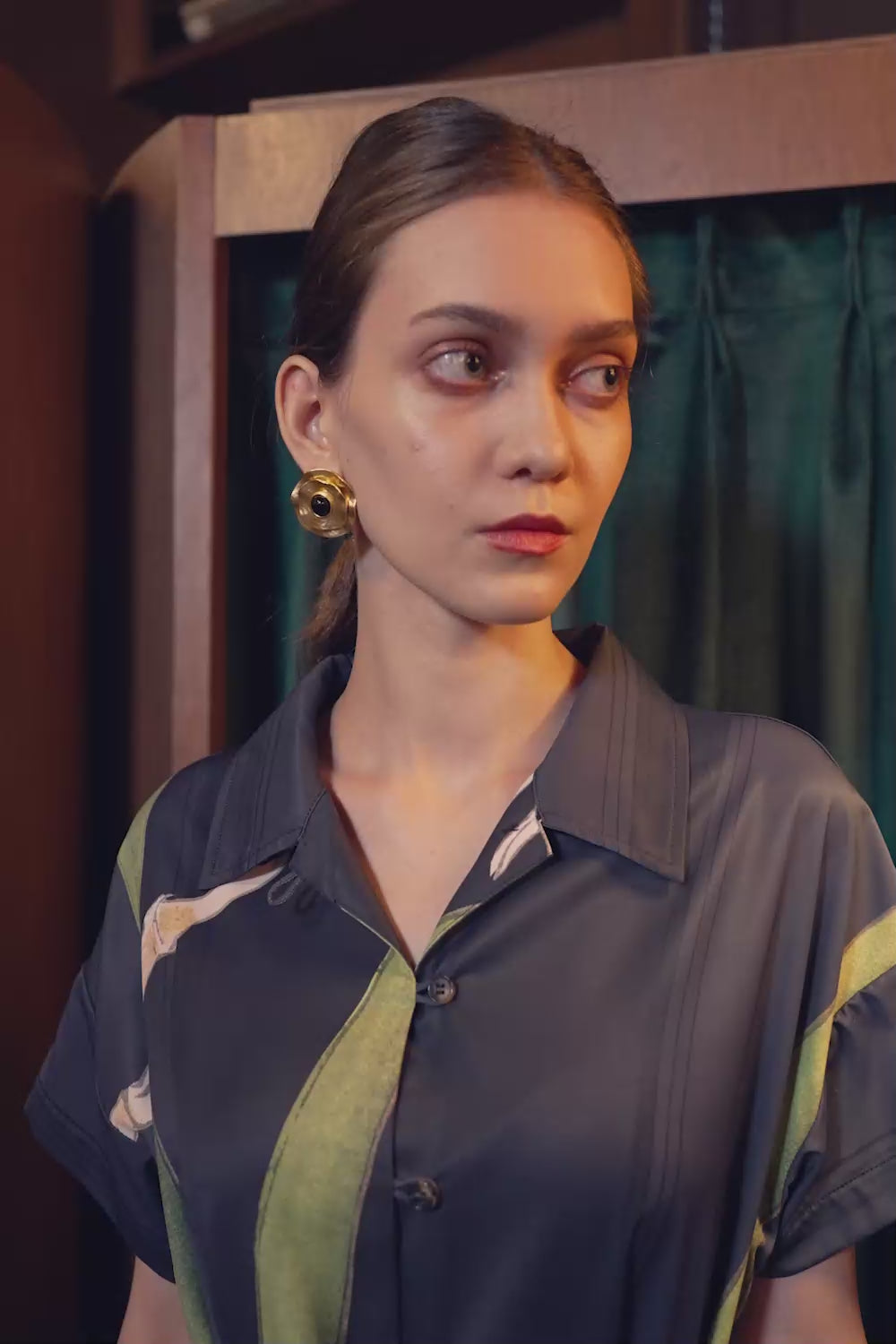 Model walking in the BEHOLD Cuban Maxi Dress, Brassavola Cucullata orchid print in black and green, showcasing button-down maxi silhouette with belted waist and floral motifs in an indoor setting.