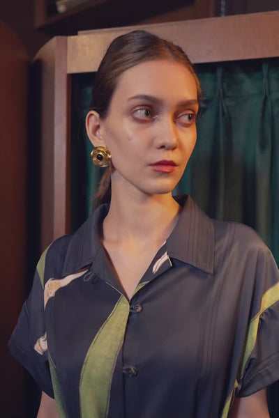 Model walking in the BEHOLD Cuban Maxi Dress, Brassavola Cucullata orchid print in black and green, showcasing button-down maxi silhouette with belted waist and floral motifs in an indoor setting.