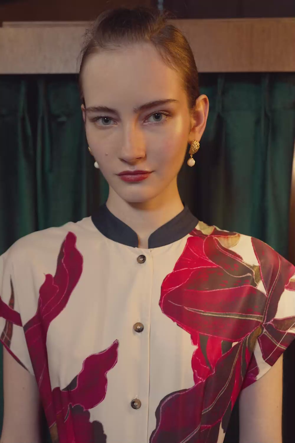 Model wearing the BEHOLD Chinese Collar Fit & Flare Maxi Dress in Laelia print, featuring crimson orchid motifs and a structured mandarin collar, standing outside a wood-panelled fitting room with emerald green curtains and warm lighting.
