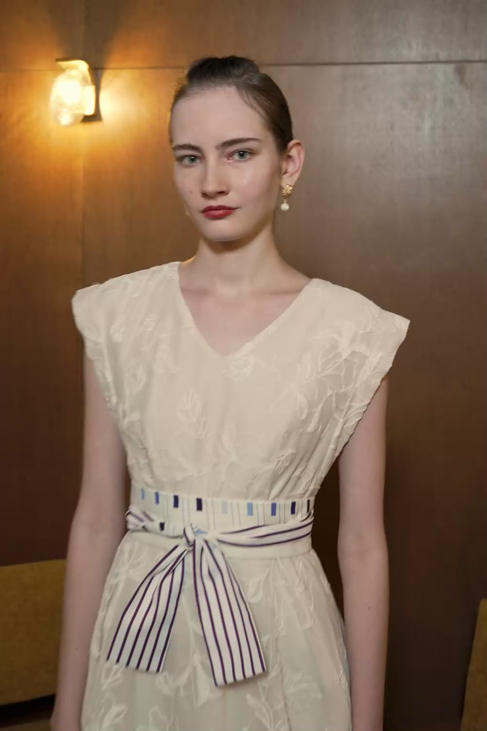Model in cream lace fit and flare dress with square sleeves and striped waist sash — elegant texture and sculptural silhouette