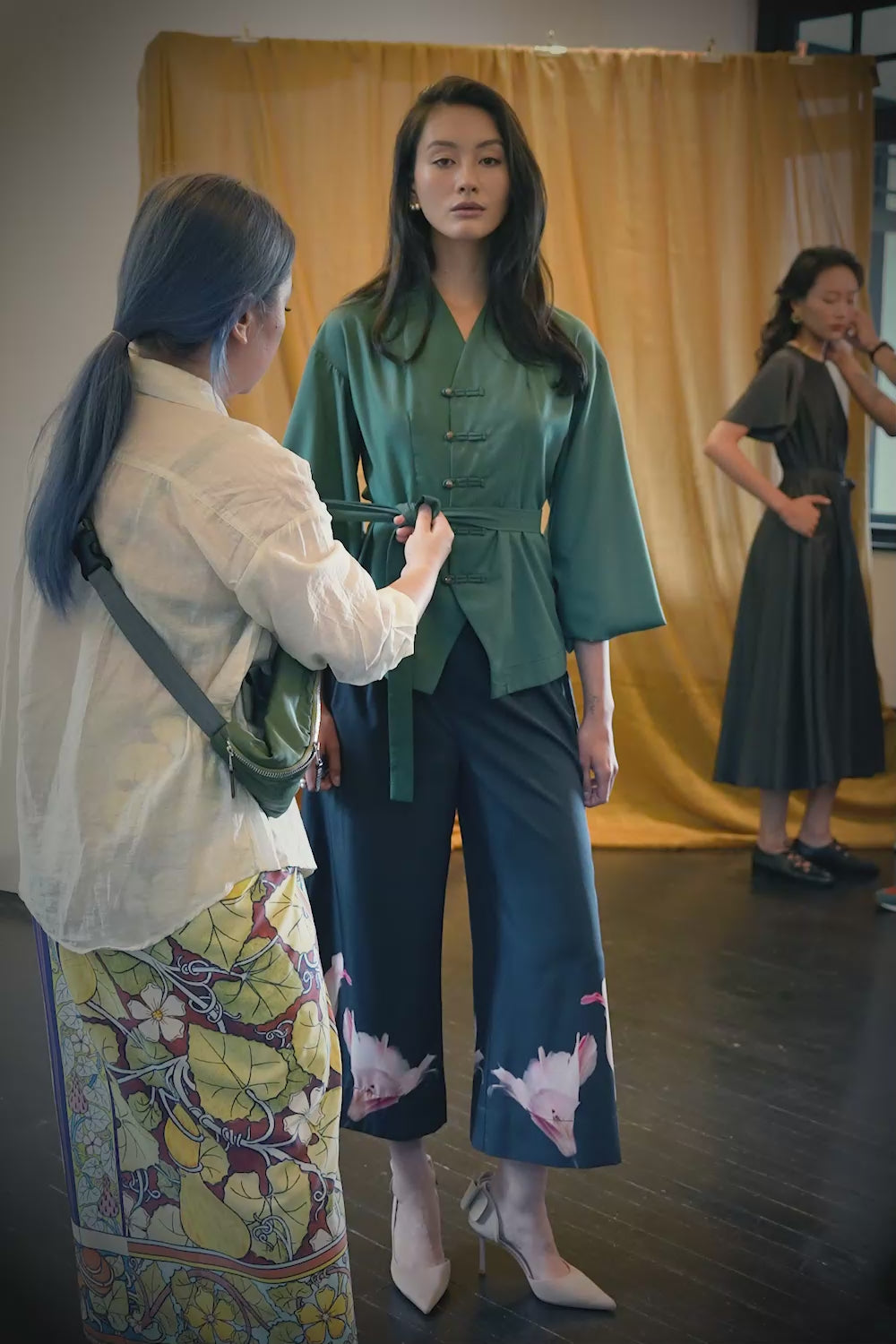 Video showing how to tie the sash on a deep green long-sleeve top with pankou button closures, featuring step-by-step waist cinching and knot styling, alongside close-up details of the top’s structured silhouette, fluid sleeves, and the dark pleated slit pants with soft drape and pink floral placement print at the hem