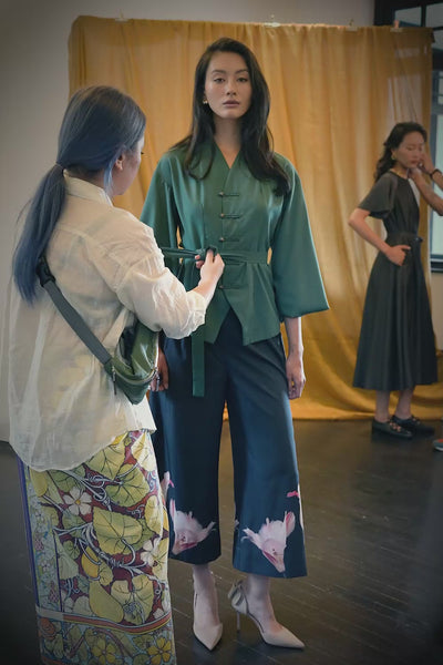Video showing how to tie the sash on a deep green long-sleeve top with pankou button closures, featuring step-by-step waist cinching and knot styling, alongside close-up details of the top’s structured silhouette, fluid sleeves, and the dark pleated slit pants with soft drape and pink floral placement print at the hem